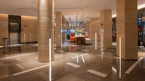 Un moderno vestíbulo de hotel con piso de mármol pulido, grandes columnas cilíndricas y una zona de estar central con sillas naranjas y una mesa.