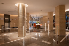 Un moderno vestíbulo de hotel con piso de mármol pulido, grandes columnas cilíndricas y una zona de estar central con sillas naranjas y una mesa.