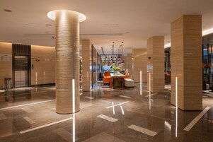 Un moderno vestíbulo de hotel con piso de mármol pulido, grandes columnas cilíndricas y una zona de estar central con sillas naranjas y una mesa.