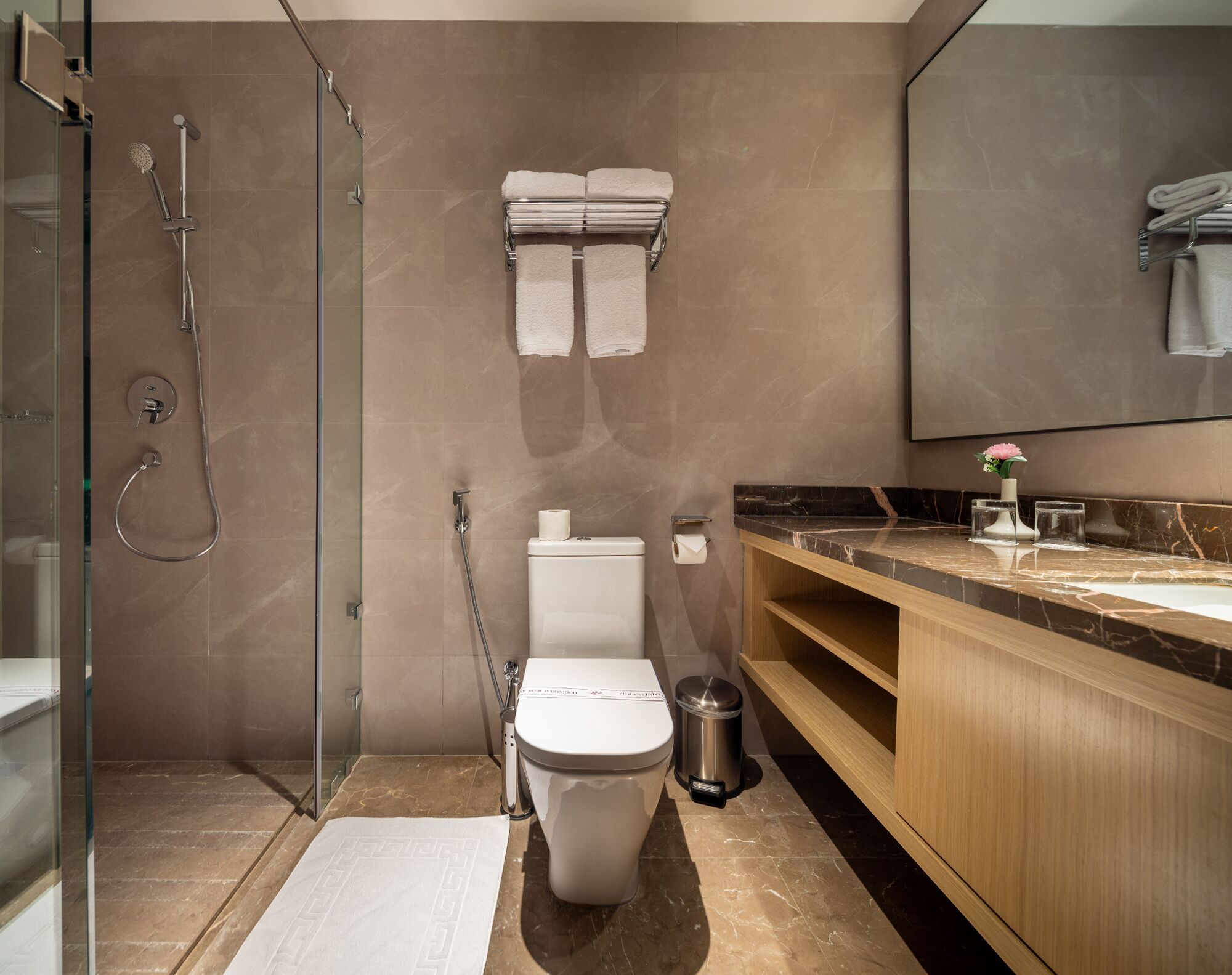 Un baño moderno con ducha envuelta en vidrio, inodoro con dispensador de toallas de papel, cubo de basura y tocador de madera con encimera de mármol.