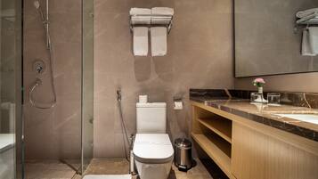 Un baño moderno con ducha envuelta en vidrio, inodoro con dispensador de toallas de papel, cubo de basura y tocador de madera con encimera de mármol.