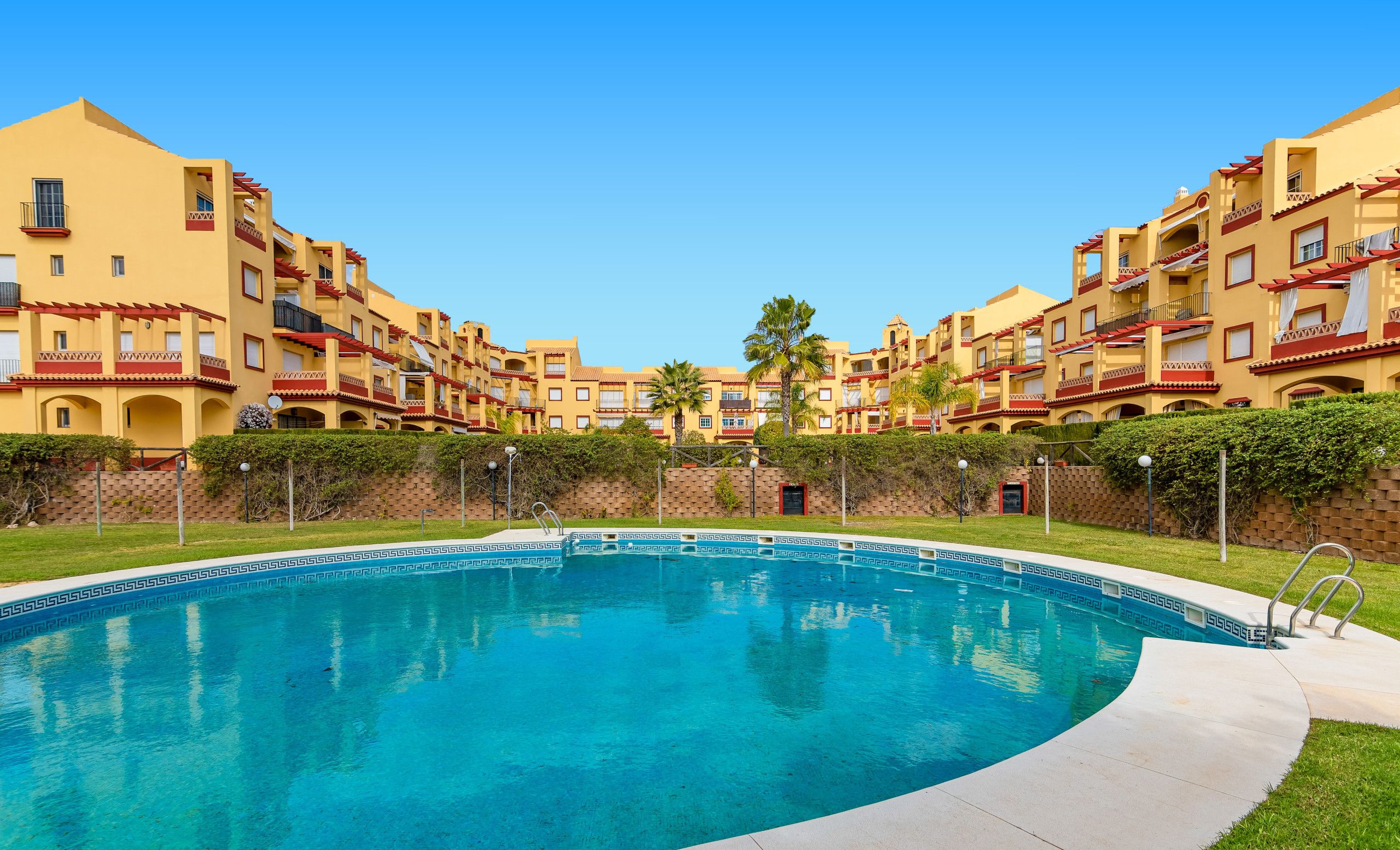 A swimming pool surrounded by multi-story buildings with balconies and a clear blue sky.