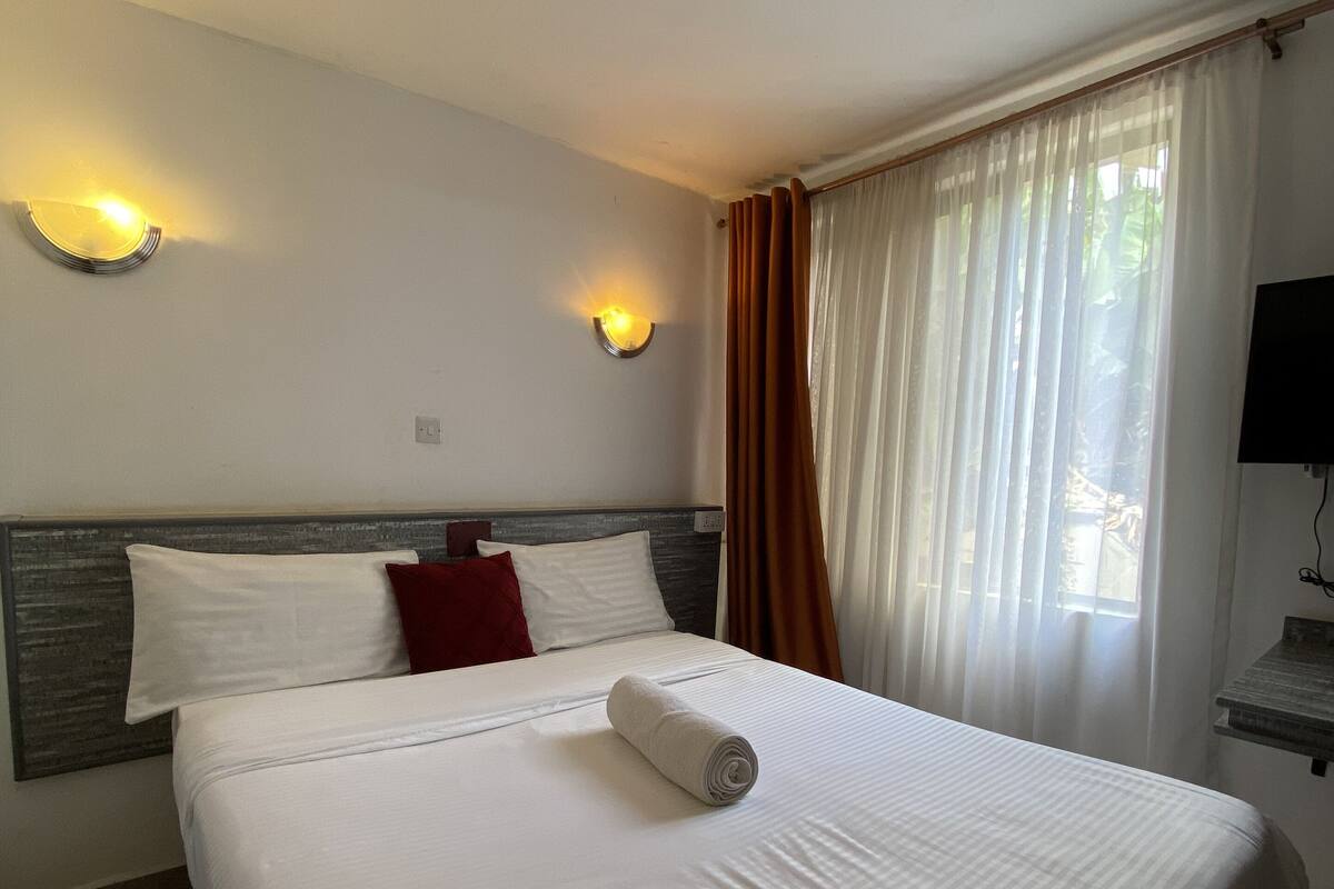 A neatly made bed with a rolled towel, a headboard, two wall-mounted lights, a TV, and a window with curtains.