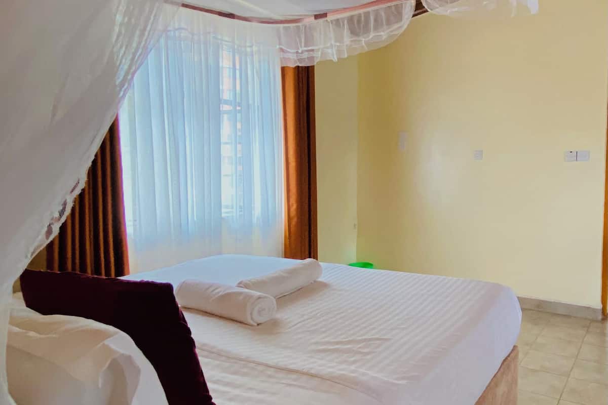 A neatly made bed with white linens and a rolled towel, a wooden headboard, and a canopy with sheer fabric.