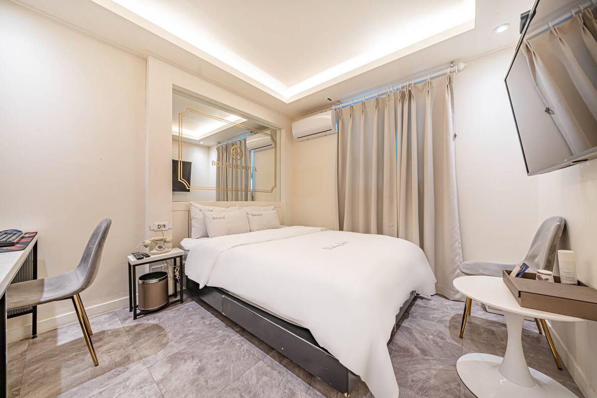 A hotel room with a bed, a desk, a chair, a small table, a trash bin, and a TV mounted on the wall.