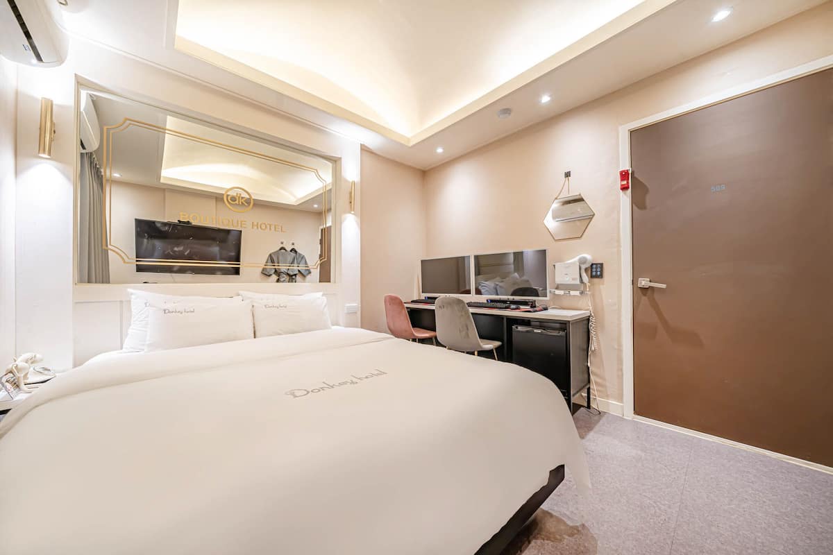 A hotel room with a large bed, a desk with a computer, a mirror, and a door with a number.