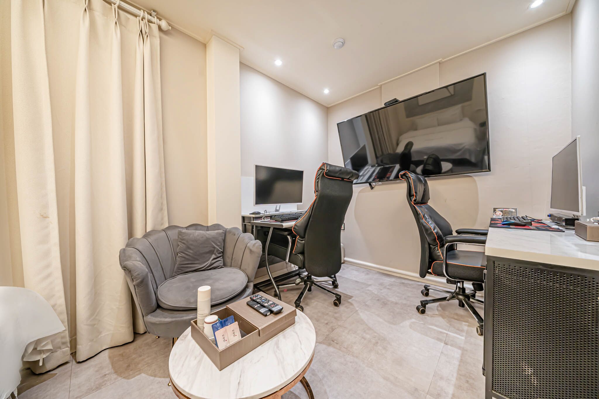 Una habitació d'hotel moderna amb un sofà gris, una taula de centre rodona amb un comandament a distància, un escriptori amb un ordinador i un gran televisor de pantalla plana fixat a la paret.
