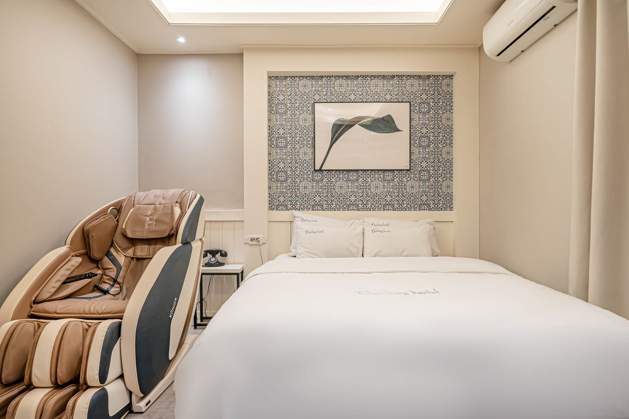 A hotel room with a massage chair, a neatly made bed, and a wall-mounted air conditioner.