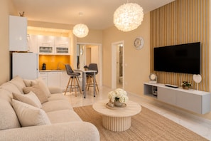 A modern living room with a sofa, coffee table, and a TV. There is a kitchen in the background with a dining area.