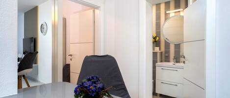 A modern bathroom with a mirror, sink, and toilet. There is a chair and a table with a vase of flowers in the adjacent room.
