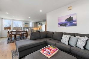 A modern living room with a grey sectional sofa, a dining table with chairs, a kitchen in the background, and a painting of a beach scene on the wall.