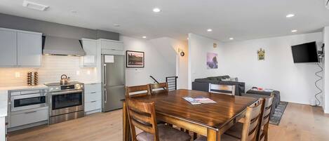A modern kitchen with stainless steel appliances, a dining area with a wooden table and chairs, and a living area with a sofa and TV.