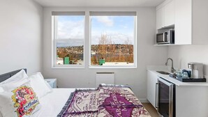 A bedroom with a bed, a view of a cityscape through two windows, a kitchenette with a microwave, and a small sink. - Hermione's Time-Turner Sweet Suite (Seattle)