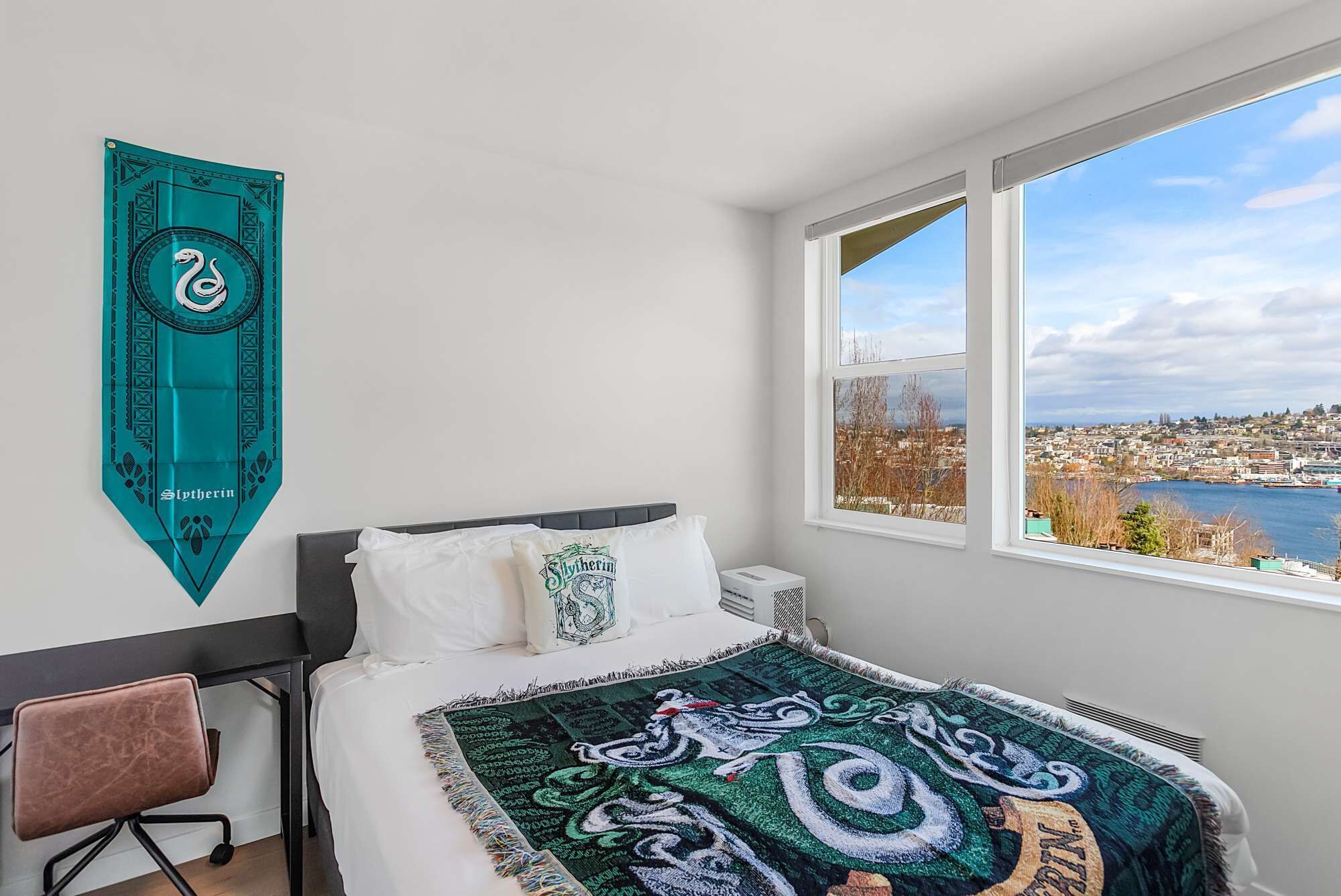 A bedroom with a bed, a chair, a window with a city view, and a teal wall hanging.