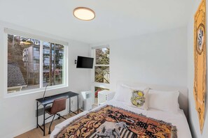 A bedroom with a bed, a desk, a chair, a TV, and a window with a view of buildings. - Hufflepuff, Scamander's Fantastic Sweet Suite (Seattle)