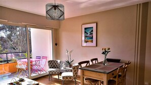 A dining area with a wooden table and chairs, a small side table with a lantern, and a view through a large window to the outside. - Port Vannes - Bretagne SUD T2 Calme Avec Terrasse (Vannes)