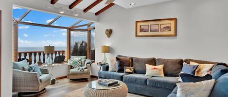 A spacious living room with a large window, a blue sofa set, wicker furniture, and a round coffee table.