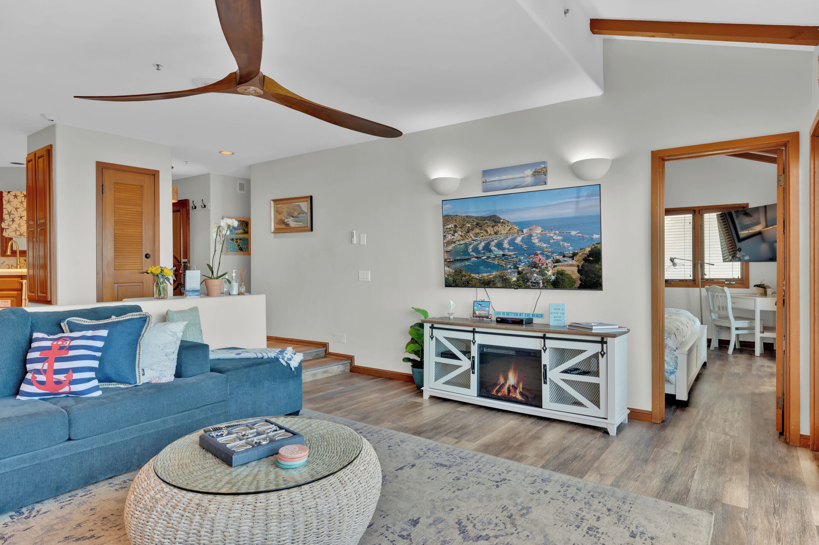A living room with a blue sofa, a fireplace, a ceiling fan, and a large painting of a coastal landscape.