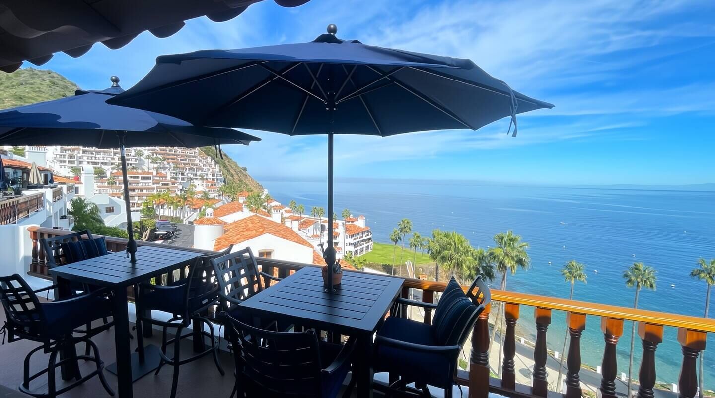 A terrace with a table and chairs, a large umbrella, and a view of the ocean and buildings.