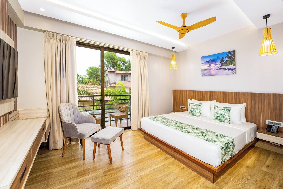 Una habitació d'hotel moderna amb un llit gran, una taula de treball, una cadira i un balcó amb vista a vegetació i edificis.