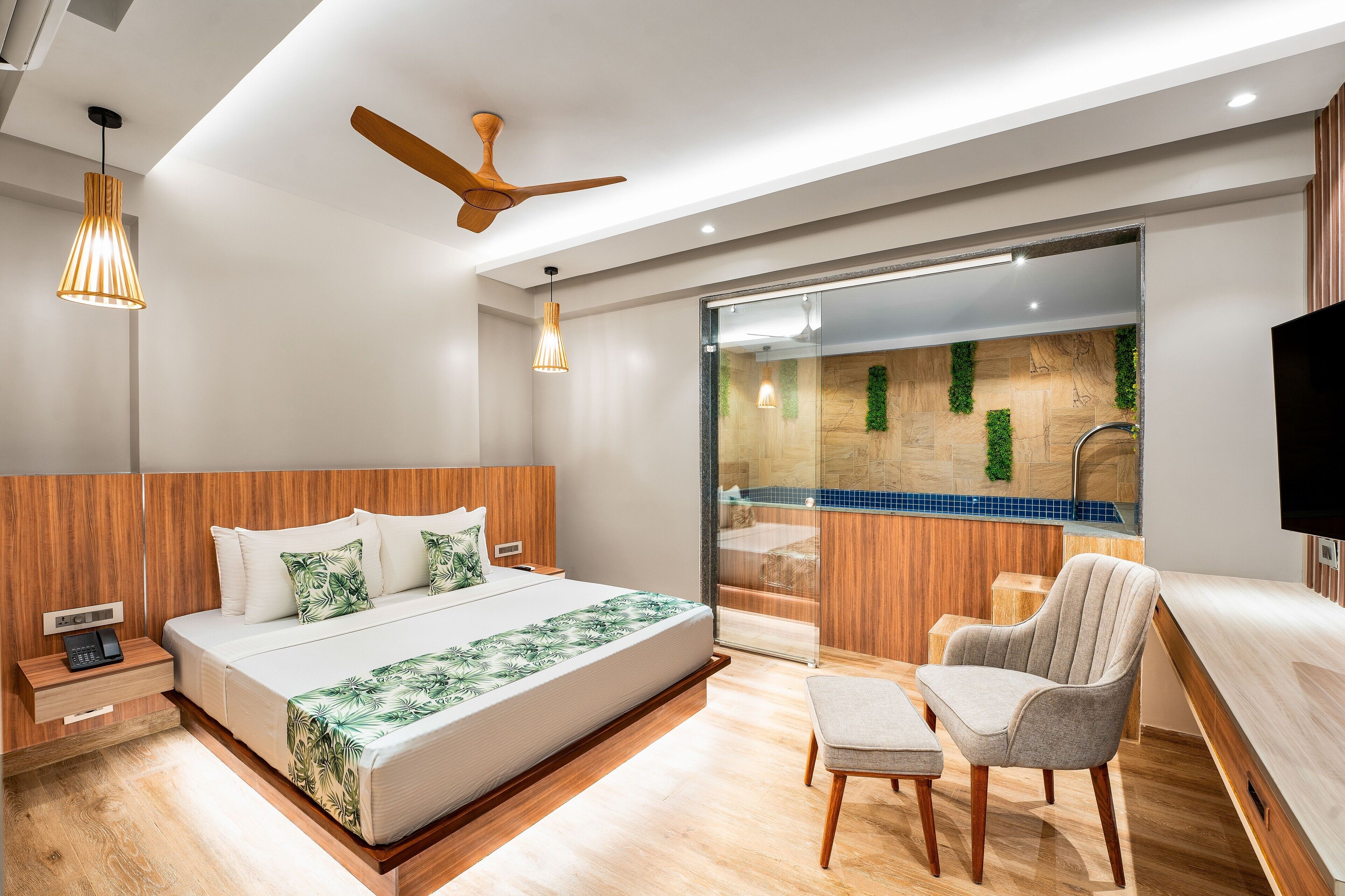 A modern bedroom with a large bed, a wooden headboard, a ceiling fan, a bedside table, a chair, and a view of a spa area through a glass door.