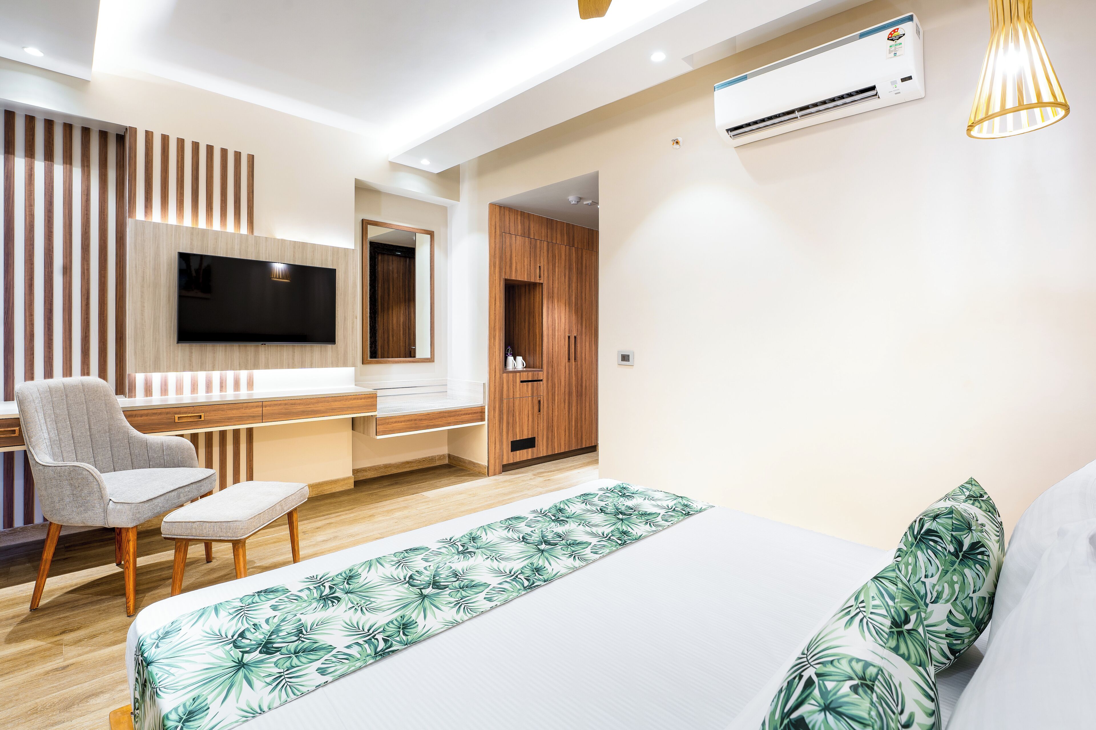 A modern hotel room with a flat-screen TV, wooden paneling, a desk, a chair, a bed with a tropical leaf-patterned bedspread, and an air conditioning unit.
