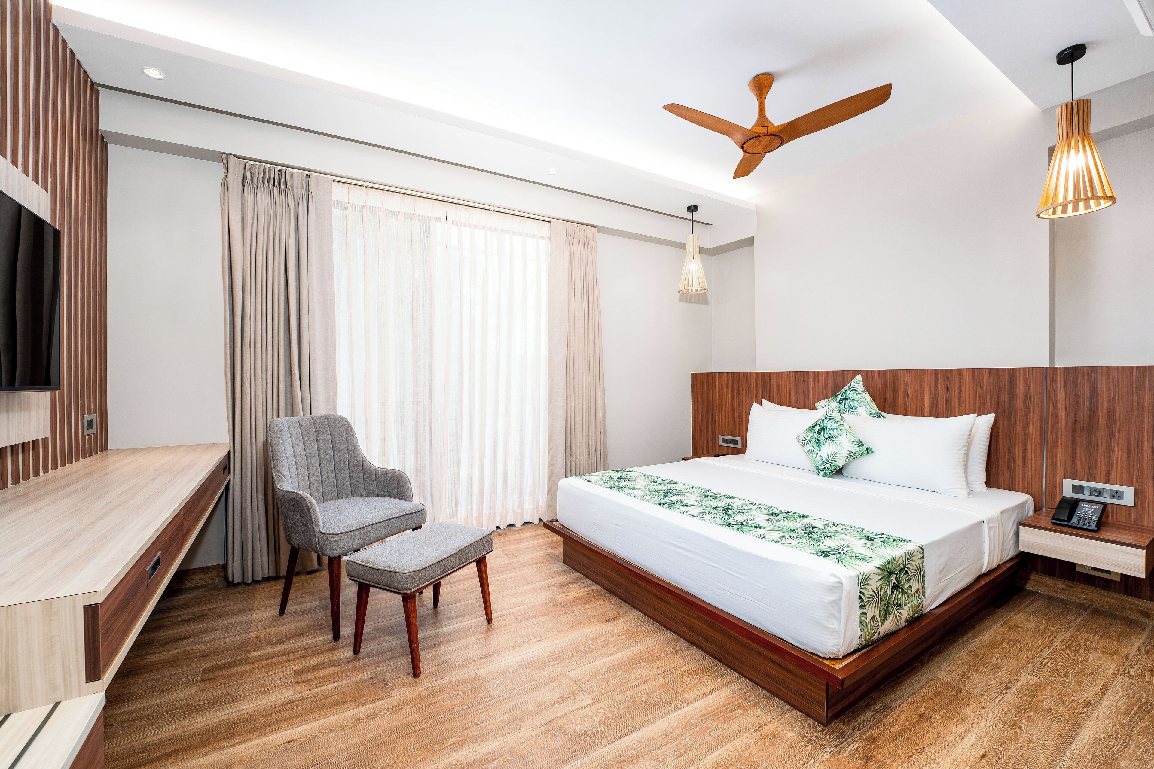 A modern hotel room with a wooden floor, a bed with a white and green floral bedspread, a grey armchair, a wooden desk, a ceiling fan, and a hanging pendant light.