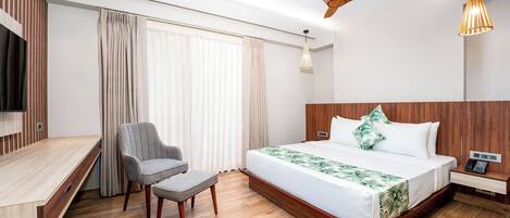 A modern hotel room with a wooden floor, a bed with a white and green floral bedspread, a grey armchair, a wooden desk, a ceiling fan, and a hanging pendant light.