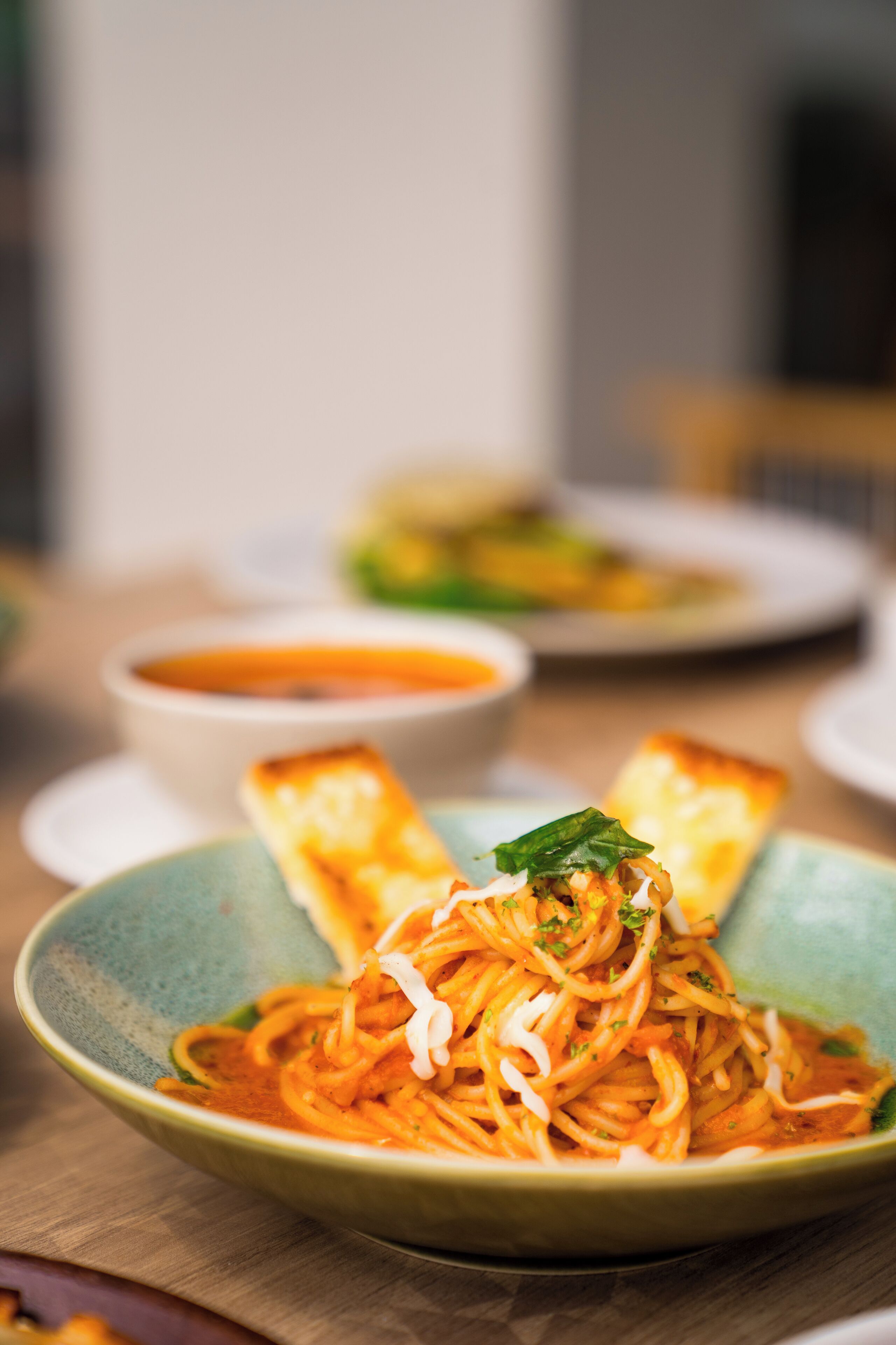 A plate of spaghetti with a garnish, a bowl of soup, and a plate with a green dish.