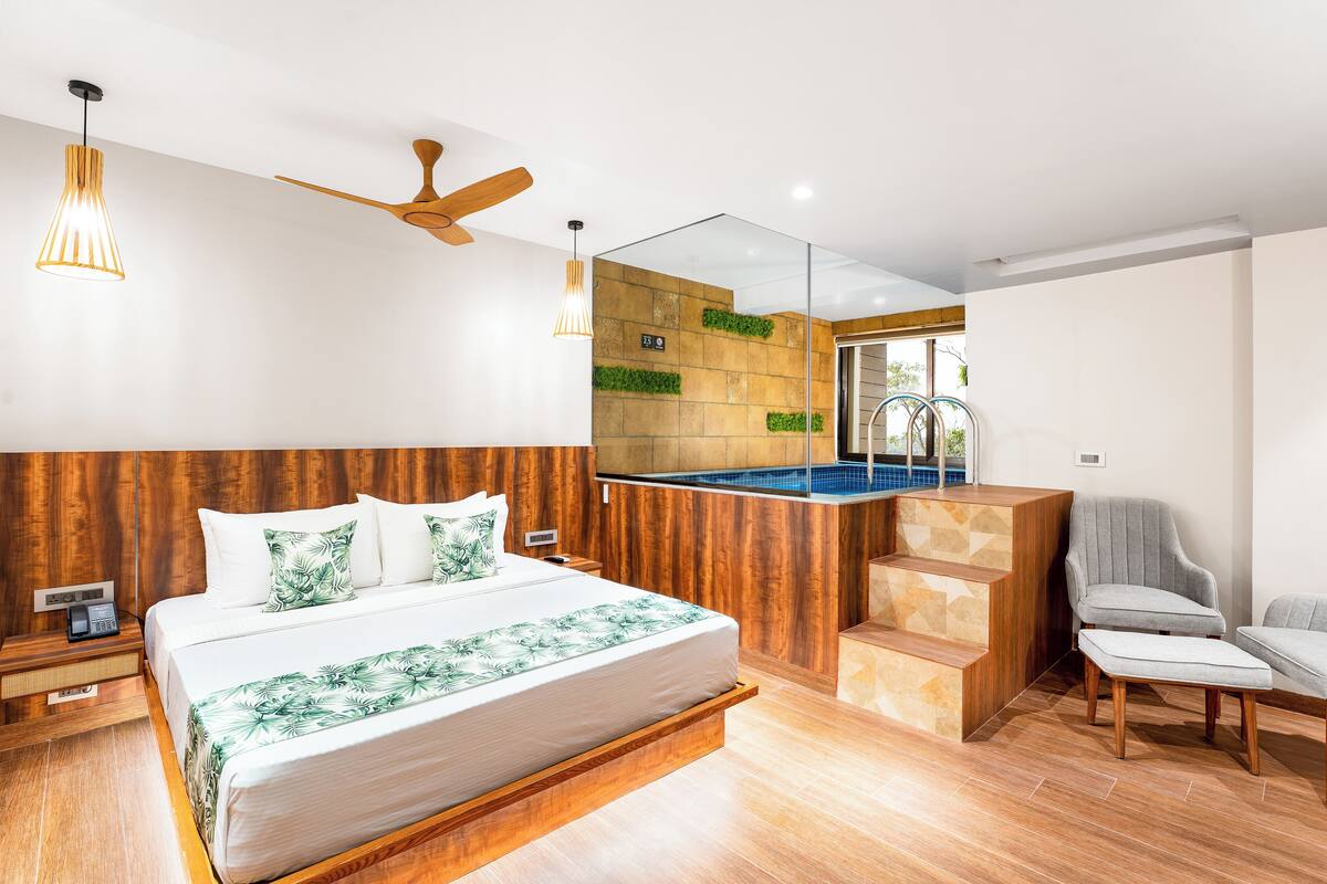 Habitació d'hotel moderna amb un capçal de llit de fusta, un llit gran amb una col·cha de fulles verdes, una tauleta de nit de fusta, un telèfon, un ventilador de sostre i una vista a la piscina a través d'una paret de vidre.