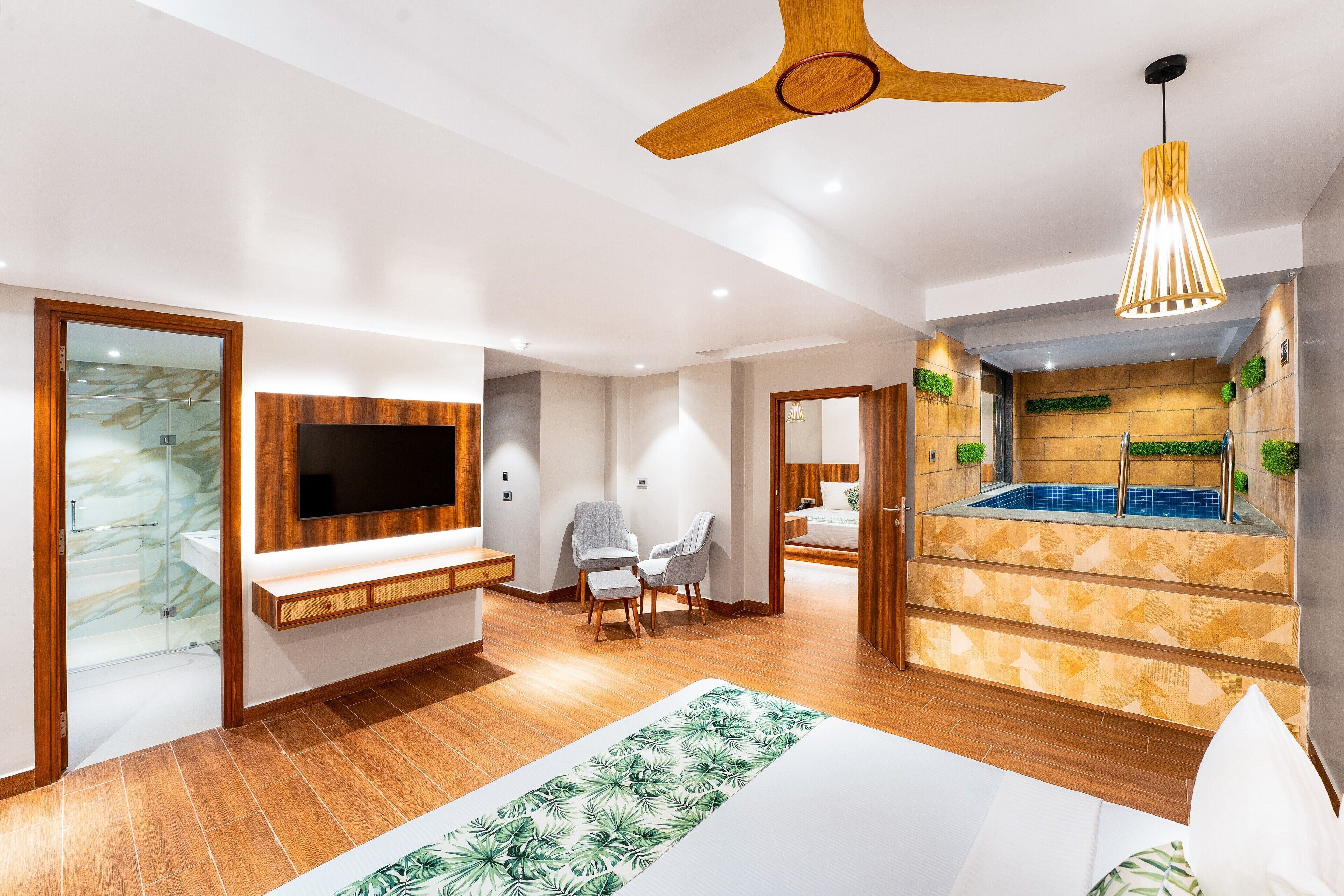 A modern hotel room with a wooden ceiling fan, a mounted TV, a seating area with chairs, a small pool, and a bed with a green leaf-patterned rug.