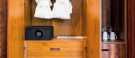 A wooden wardrobe with two white robes hanging, a black safe, and a pair of slippers on the lower shelf.