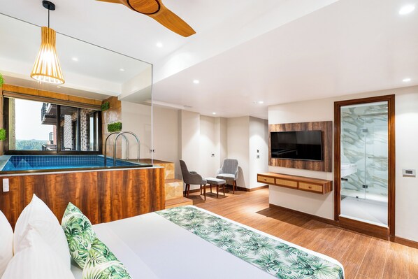 A modern hotel room with a large bed, a desk, a chair, a TV, and a small pool area.