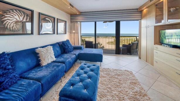 A modern living room with a blue sofa, a television, and a balcony with a view of the ocean.