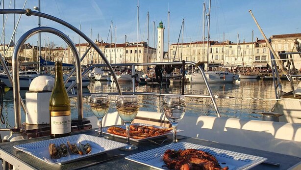 Ein Esstisch mit Meeresfrüchten, Weingläsern und einer Flasche auf einem Boot in einer Marina, im Hintergrund weitere Boote und Gebäude.