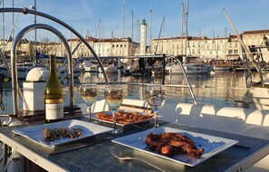 A dining table with seafood dishes, wine glasses, and a bottle on a boat in a marina with other boats and buildings in the background. - Nuits au Port - Grand Voilier à Quai - Coriandre II (La Rochelle)
