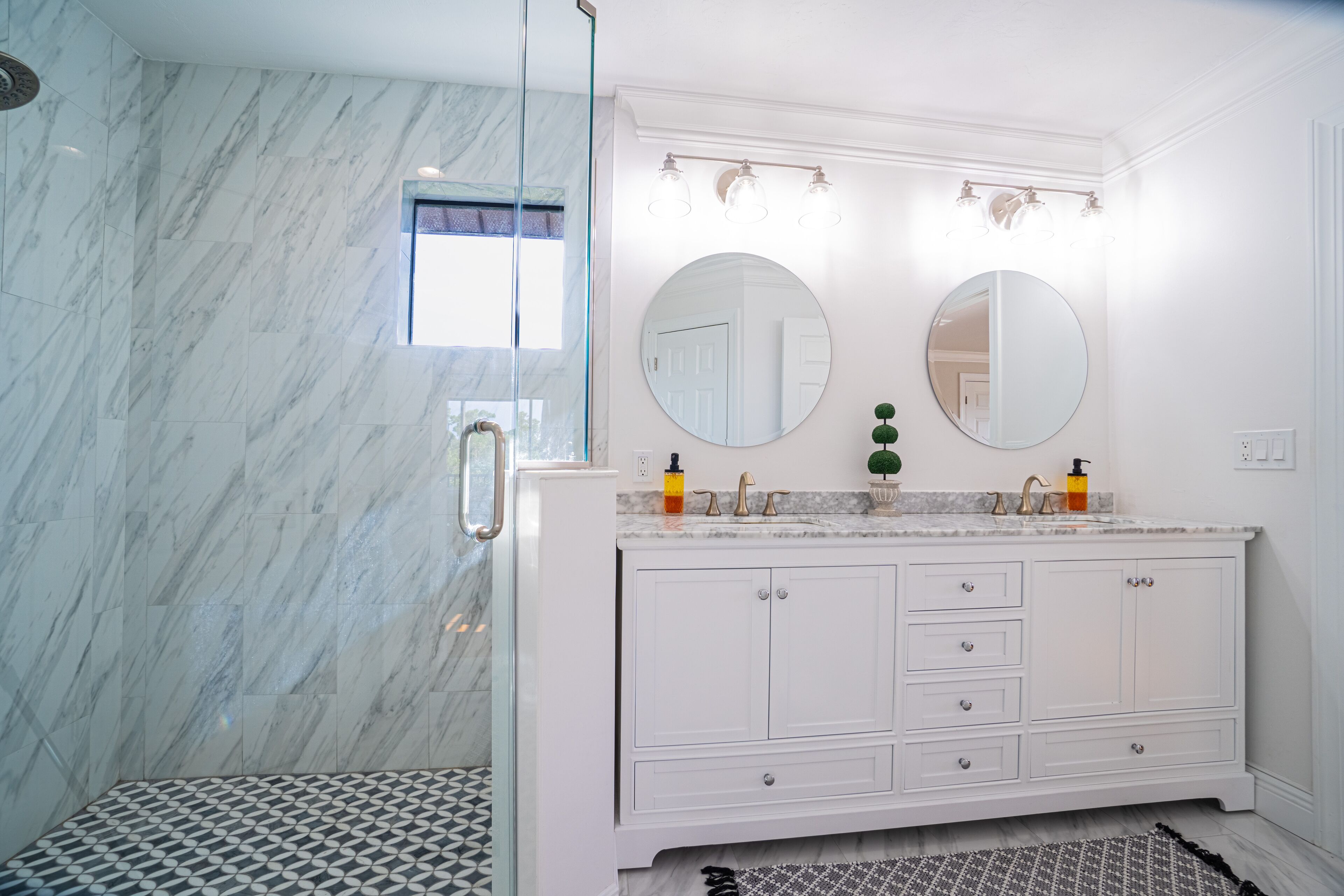 Baño con pared de mármol, cabina de ducha de vidrio, tocador blanco con dos lavamanos y dos espejos redondos.