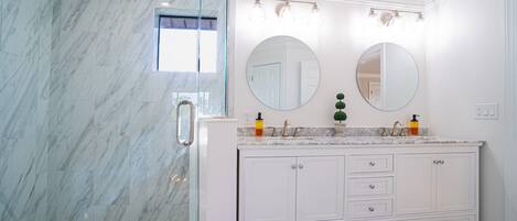 Baño con pared de mármol, cabina de ducha de vidrio, tocador blanco con dos lavamanos y dos espejos redondos.