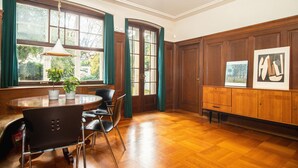 Un coin repas avec une table en bois, des chaises et un luminaire suspendu. Il y a deux fenêtres avec des rideaux, un meuble contenant des œuvres d’art et une plante en pot sur la table.