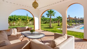 A spacious patio with a round table and beige sofas, large arched windows, and a view of a pool and palm trees. - Villa Leila Farmhouse  (marrakech)