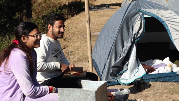 Dos personas sentadas en una mesa al aire libre, con una tienda de campaña al fondo.