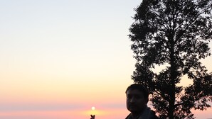 Una persona de pie junto a un árbol, con un atardecer de fondo.