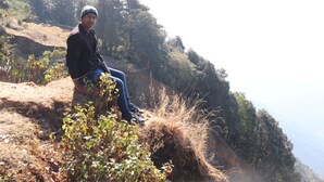 Una persona sentada en una ladera cubierta de hierba, con árboles al fondo.