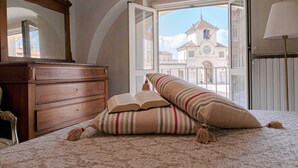 A bedroom with a bed, a wooden dresser, and a large window with a view of a building. - ROYAL HOUSE on the Palace of Venaria (Venaria Reale)