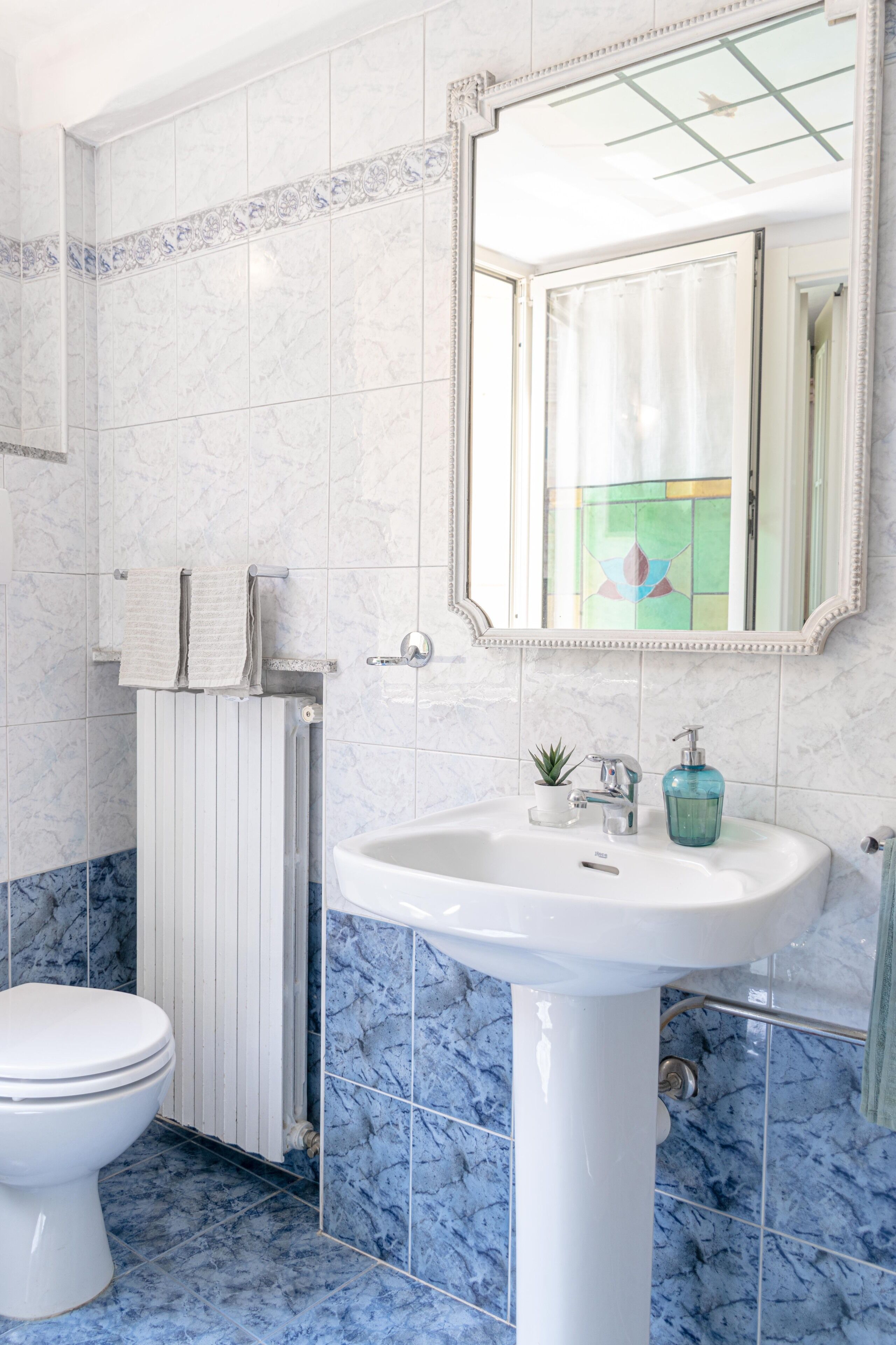 Une salle de bain avec des toilettes blanches, un lavabo blanc doté d’un distributeur de savon en verre bleu, et un miroir avec un cadre décoratif.