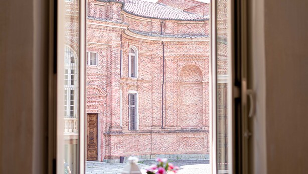 A view through a window of a brick building with arched windows and a door, a potted plant, and a candle on the windowsill. - ROYAL HOUSE on the Palace of Venaria (Venaria Reale)