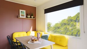 Una habitación con un sofá amarillo, una mesa con una bandeja de pasteles, un cuadro en la pared y una ventana con vistas a árboles.