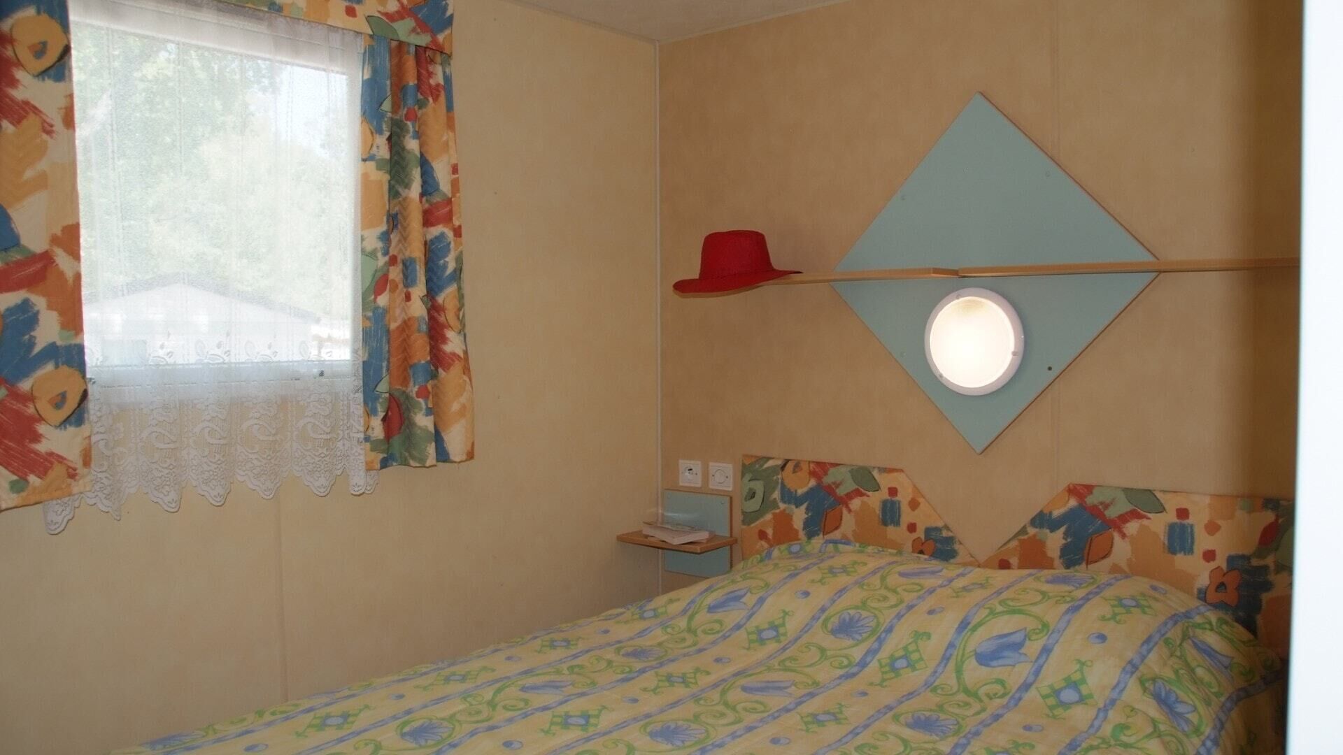 Une chambre à coucher avec un lit, une fenêtre avec des rideaux, un miroir fixé au mur et une petite table de chevet.