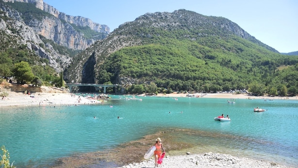 Una playa con agua turquesa cristalina, terreno rocoso y un puente al fondo.