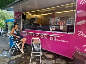 Un camion-restaurant à la façade rose, avec un panneau affichant le menu et une personne assise à une table à l’extérieur.